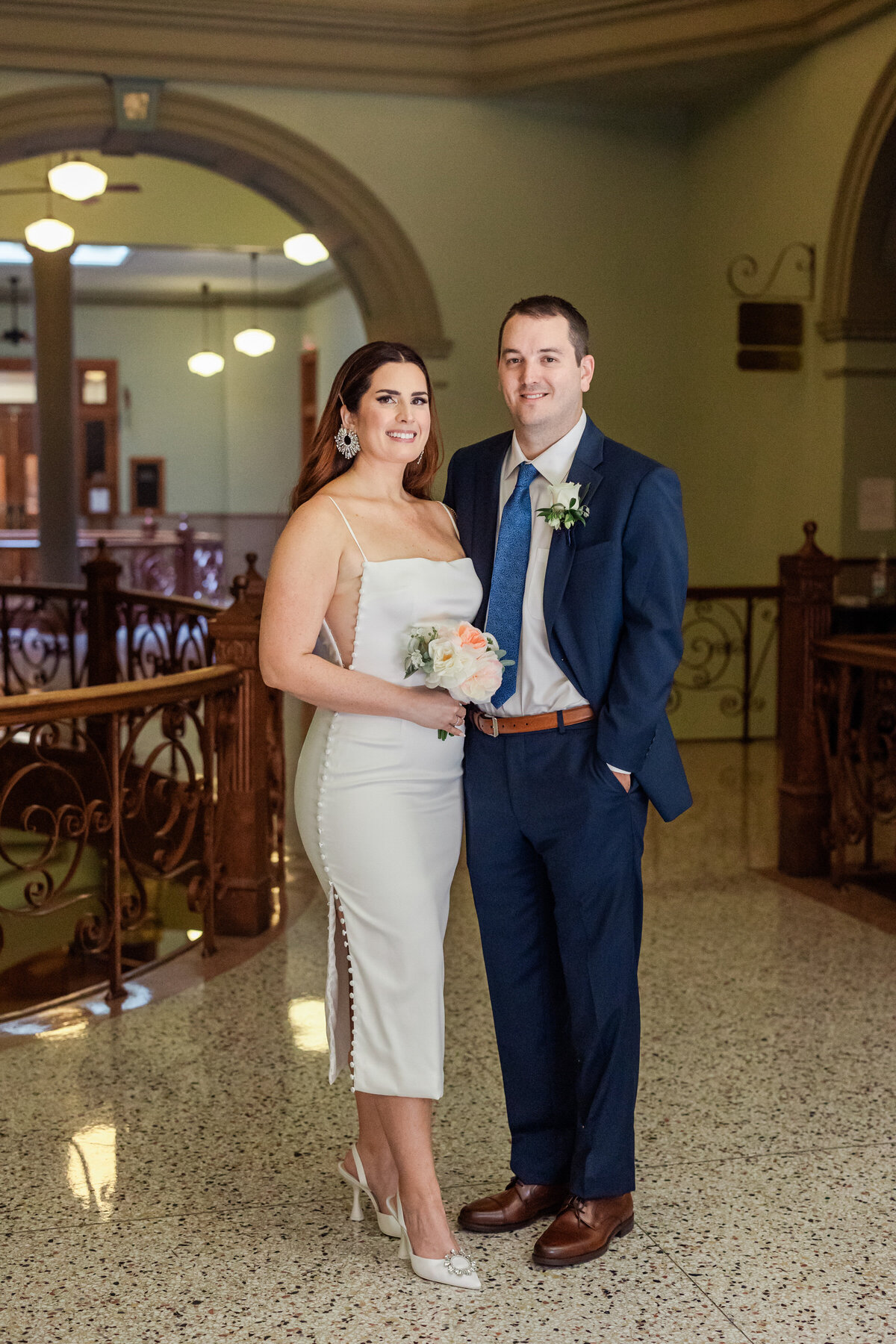 Portrait of a bride and groom after their elopement ceremony at the Tarrant County Courthouse in Fort Worth, Texas. The bride is on the left and is wearing a simple yet stylish white dress and is holding a bouquet. The groom is on the right an dis wearing a navy suit with a boutonniere.