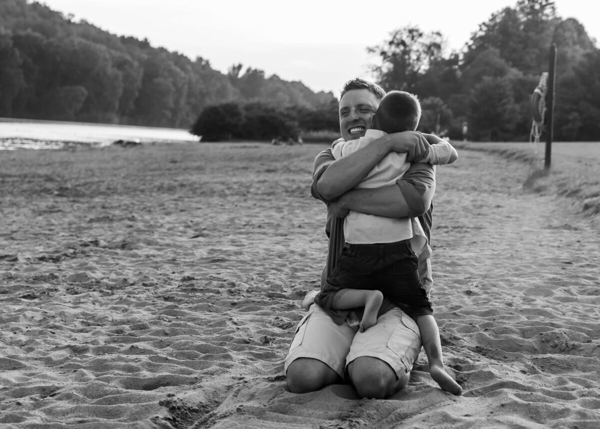 Black and white photo of a man embracing a child on the beach, captured by a Pittsburgh maternity photographer.