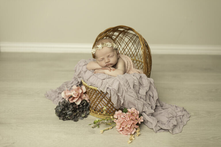 Newborn baby girl peacefully sleeping in a wicker basket, surrounded by delicate flowers, creating a soft and dreamy floral portrait.