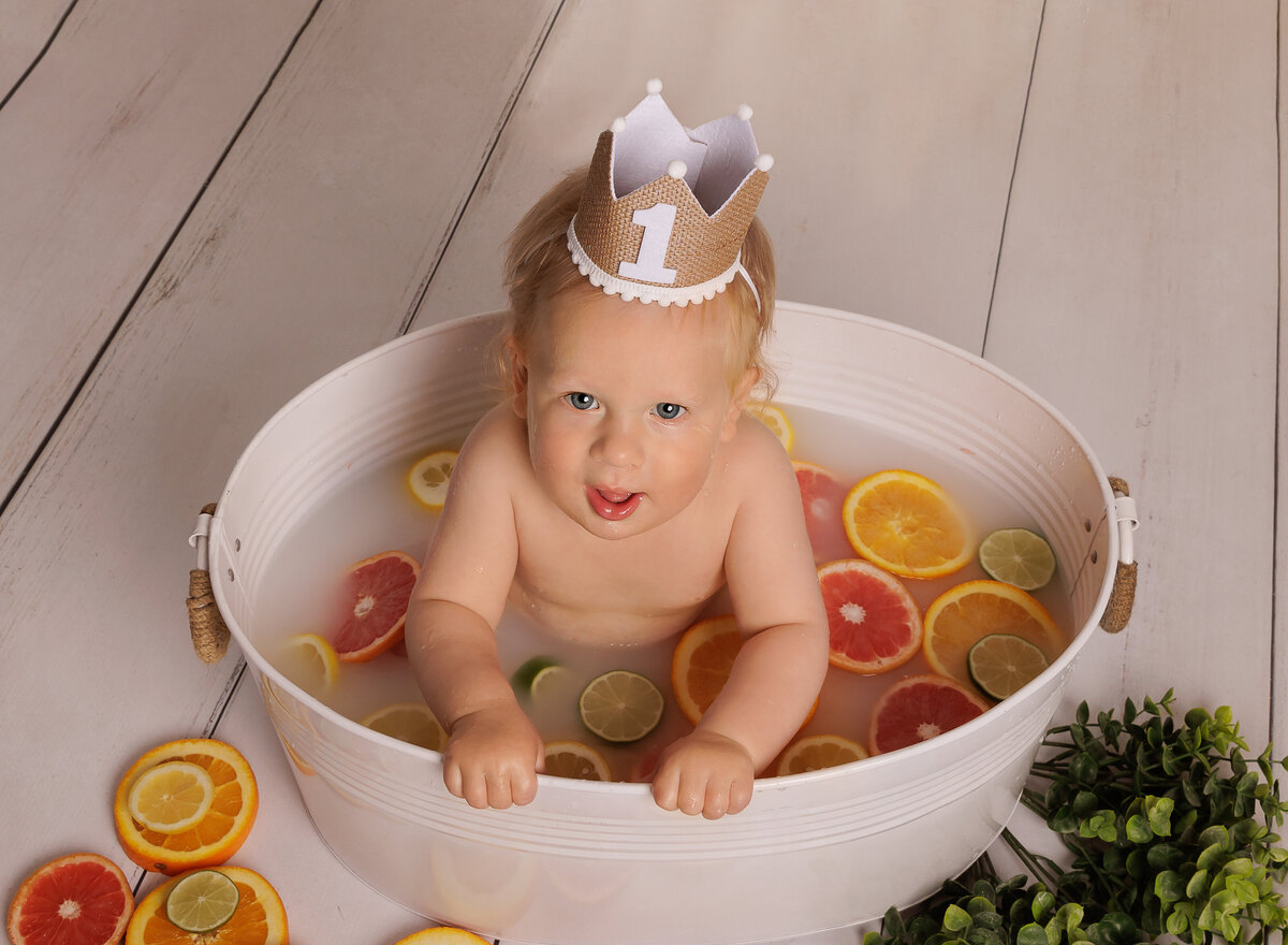 Baby boy is in a meal wash basin for a bath after his first birthday cake smash. The water is filled with sliced oranges and grapefruit. Baby is looking up at the camera and smiling. Captured by Chaya Bornstein brooklyn milestone photographer