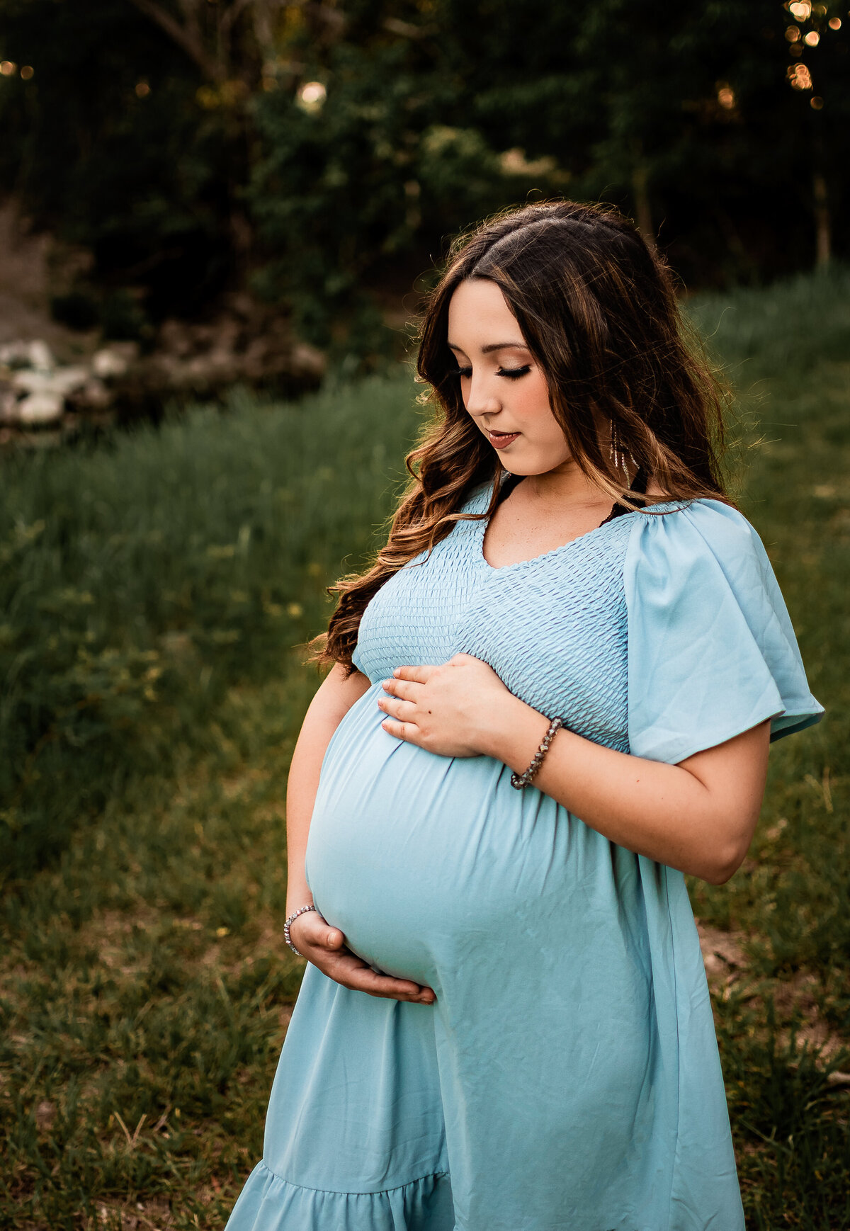 At a Brazoria county park, a mom to be wearing a blue dress holds her belly.
