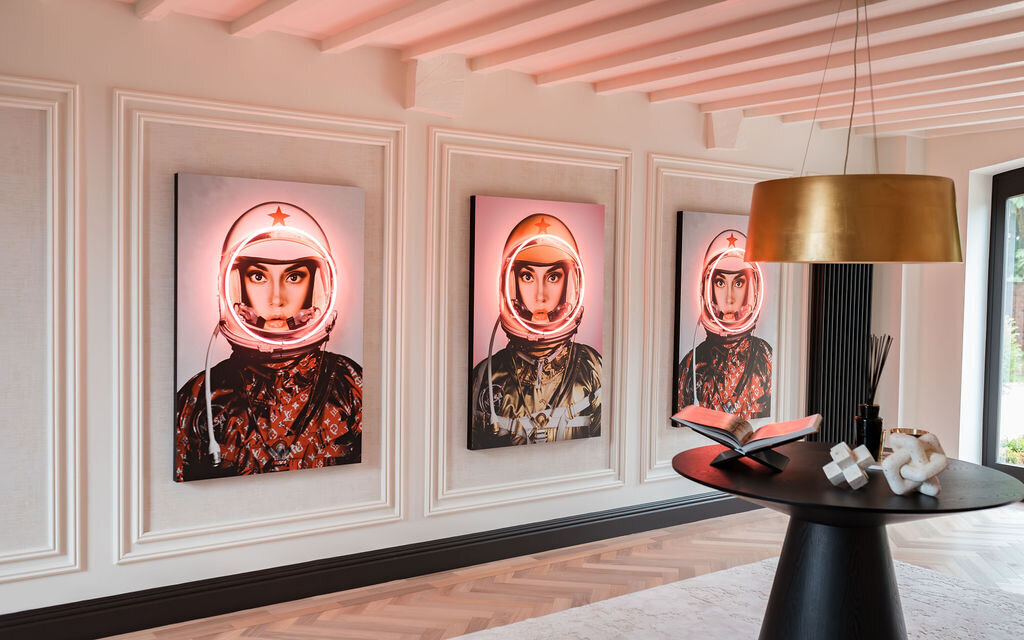 A modern interior features three portraits of astronauts with white helmets illuminated by pink LED, framed on a white paneled wall. Centered is a black round table with decorative objects beneath a gold ceiling light. Wooden floorboards and a window add to the aesthetic.