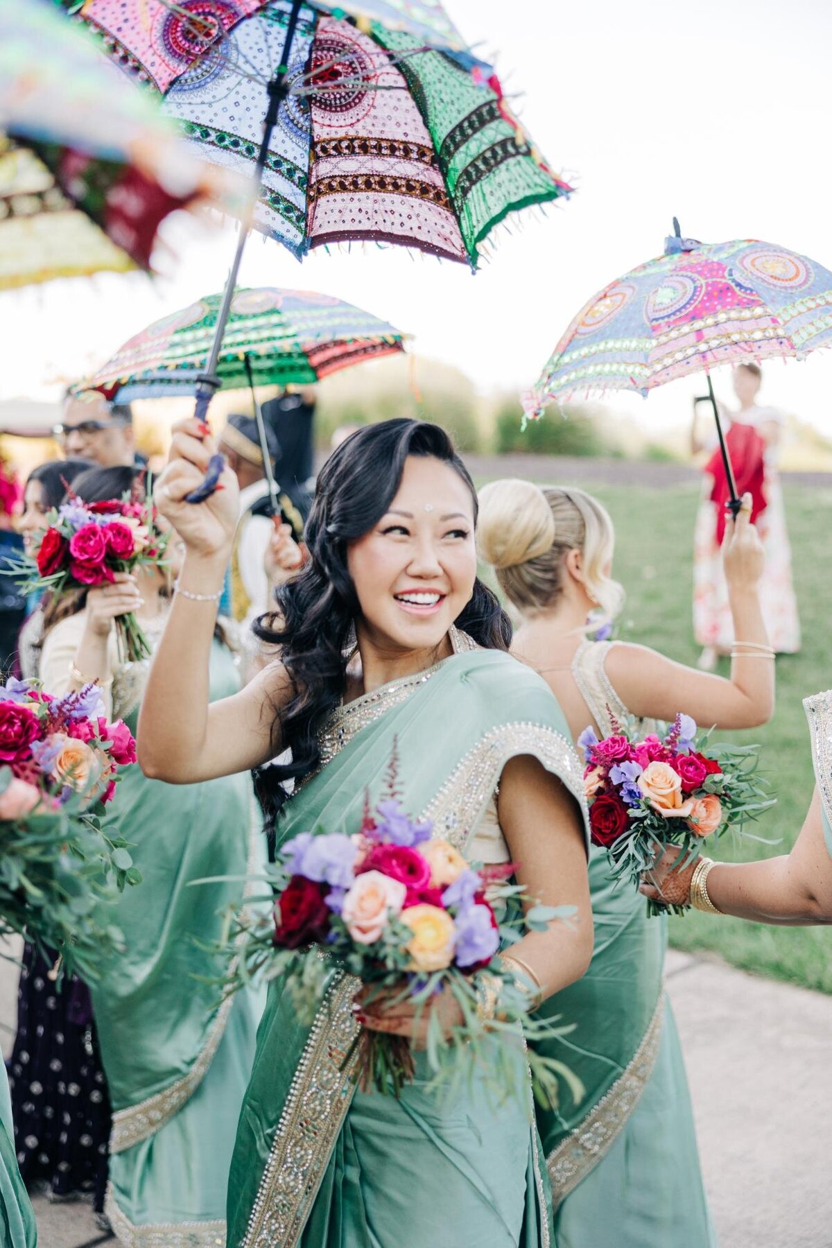 A woman in a green sari smiles while holding a bouquet, surrounded by others with colorful parasols.