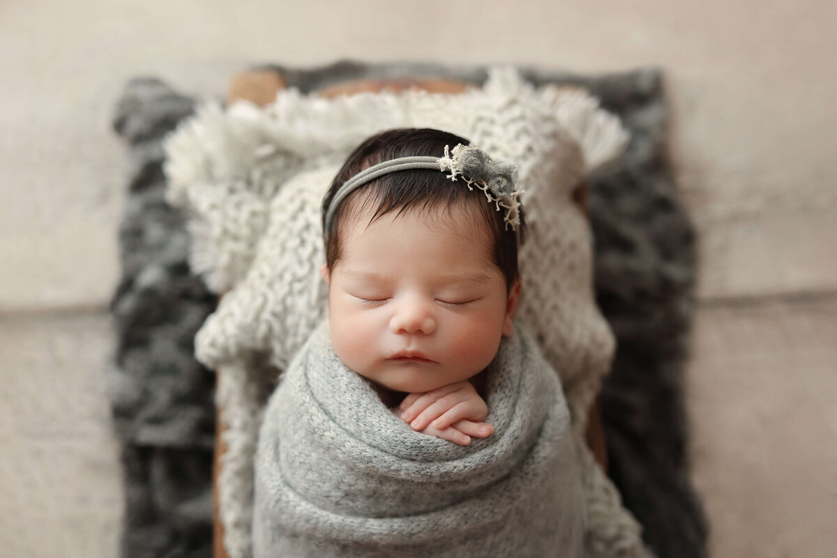 baby girl in a grey knit wrap and headband laying in a wooden bowl on a white wood floor in a newborn session