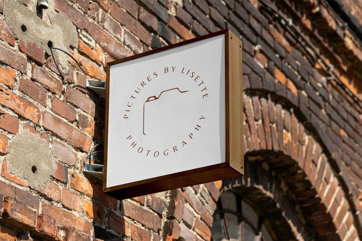 Wooden-framed square sign mounted on a rustic red brick wall, displaying the circular logo “Pictures by Lisette Photography” with a minimalist camera outline.