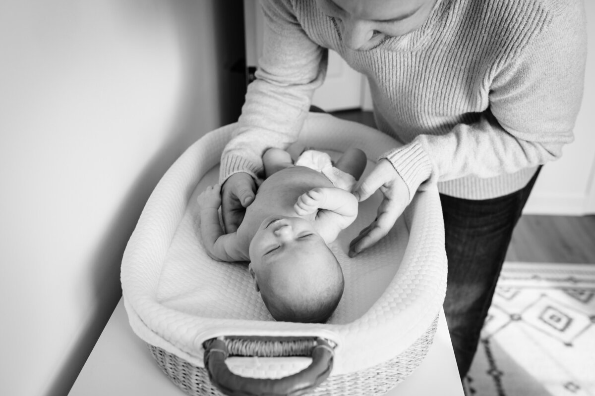 A black-and-white photo of a father gently touching his newborn baby, who is lying in a basket on a changing table. The baby, dressed only in a diaper, is calmly looking up as the father smiles down.