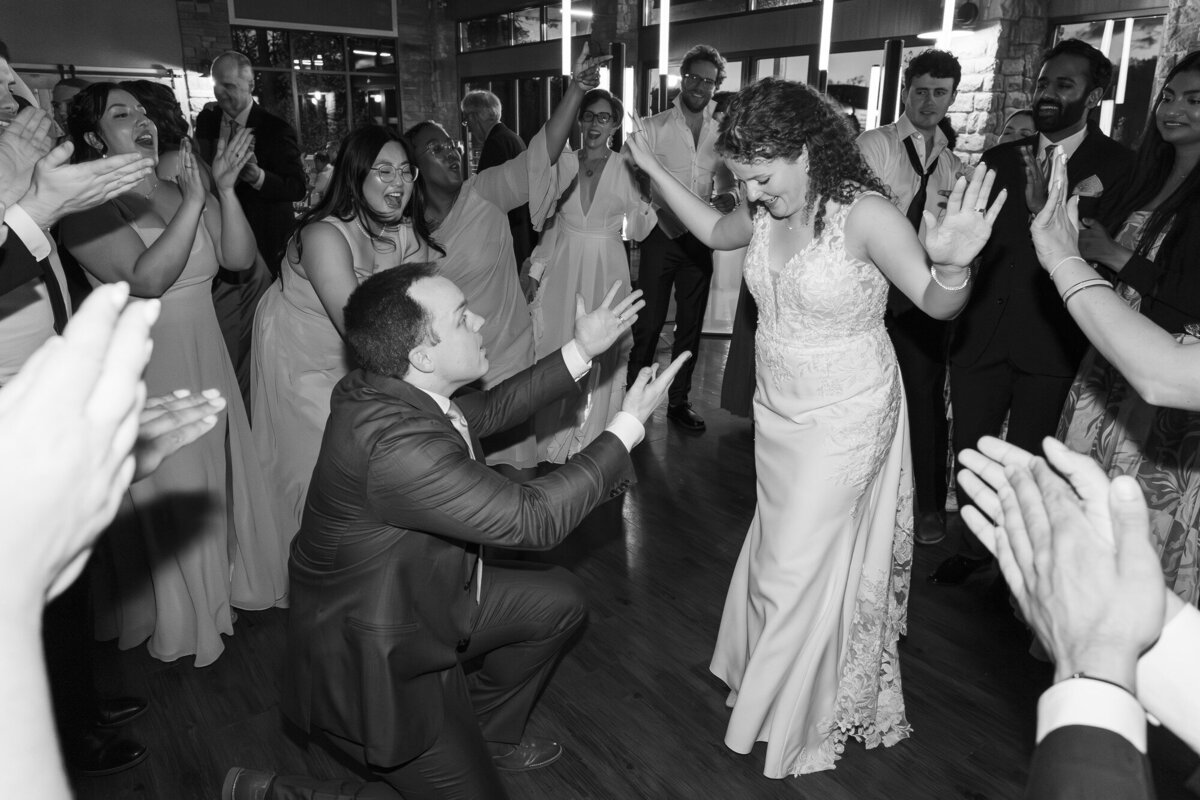 Black and white dance floor photo of guests cheering around a bride and a kneeling groom