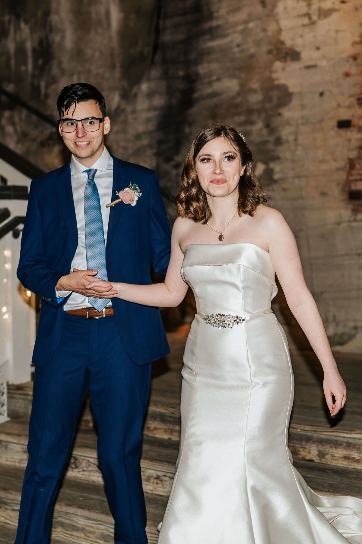 A bride in a strapless white gown and a groom in a blue suit with a pink boutonniere stand together, holding hands, smiling and posing indoors against a brick wall.