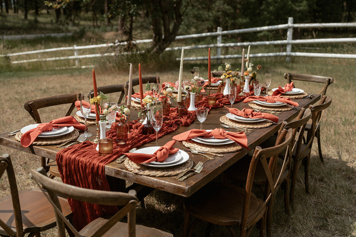 A rustic outdoor elopement table styled with terracotta napkins, amber glassware, woven placemats, and floral centerpieces for an intimate dinner in the woods.