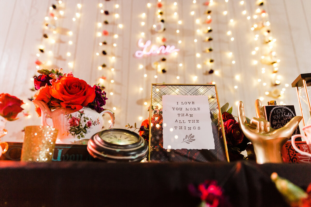 Detail shot of a wedding centerpiece display at a wedding reception in Denton, Texas. The table has a black tablecloth and is covered with an array of items: flowers in vases, clocks, a gold hand statue making a "Love" gesture, a sign reading, "I love you more than all the things," and other knickknacks. In the background are strings of lights hanging on the wall alongside a neon cursive sign reading "Love."