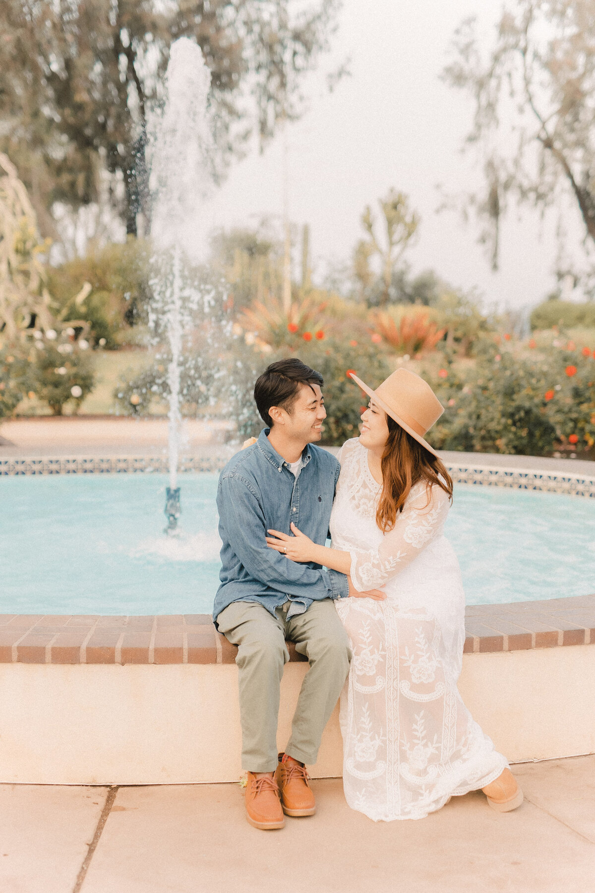 Romantic maternity photo at Balboa Park in San Diego, featuring an expecting couple sitting by a fountain, with the mother in a lace white dress and tan hat.