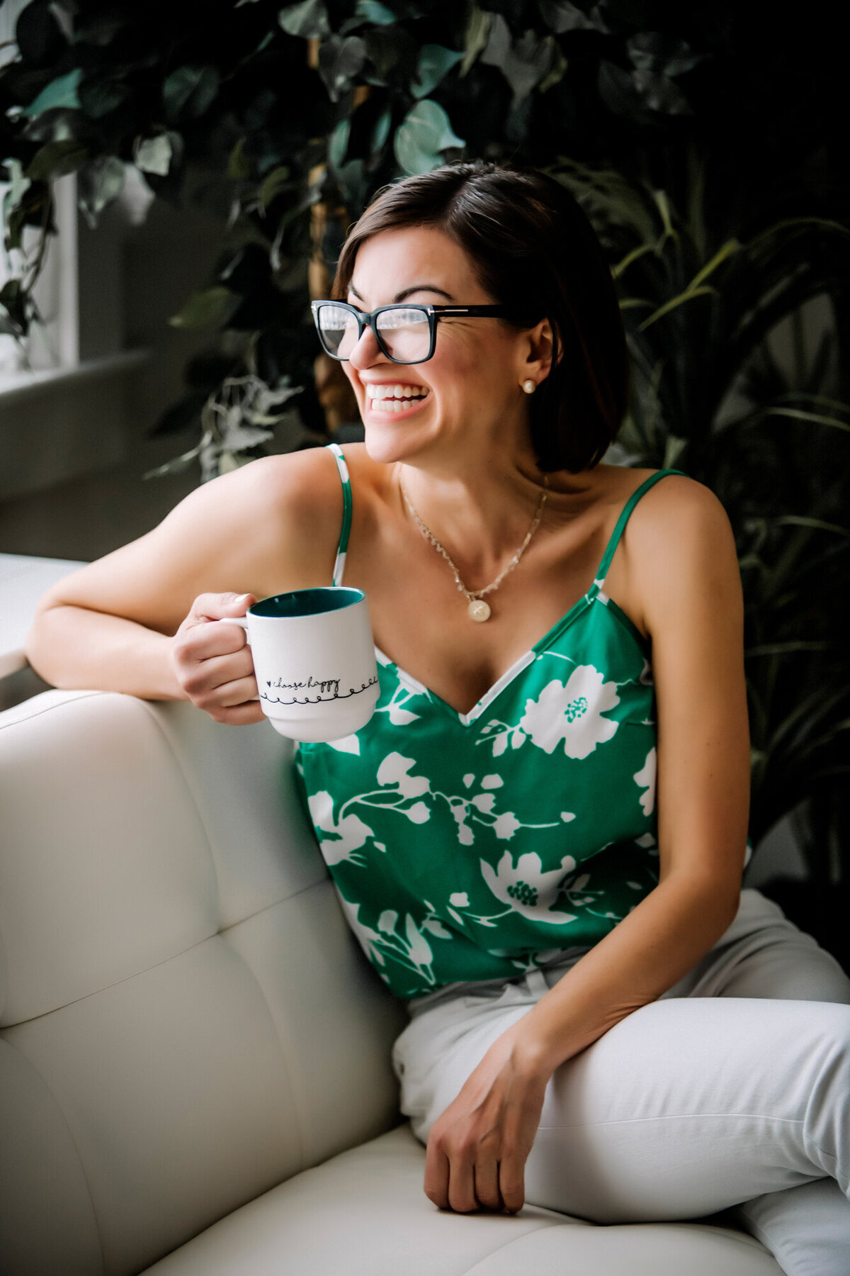 A female dating portrait client wearing a green shirt with white flowers, sitting on a white couch, holding a cup of coffee while looking out the window