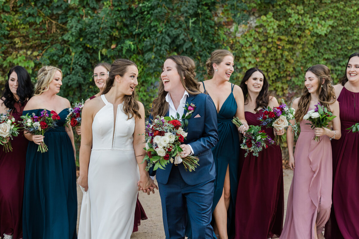 Portrait of two brides and their wedding party at laughing and standing together before their wedding ceremony at the BRIK Venue in Fort Worth, Texas. The bride on the let is wearing a sleeveless, elegant, white dress while the bride on the right is wearing a navy suit with a boutonniere and is holding a bouquet. The wedding party members are all wearing dresses in shades of burgundy, pink, or blue and are all holding bouquets in front of a large wall covered in greenery.
