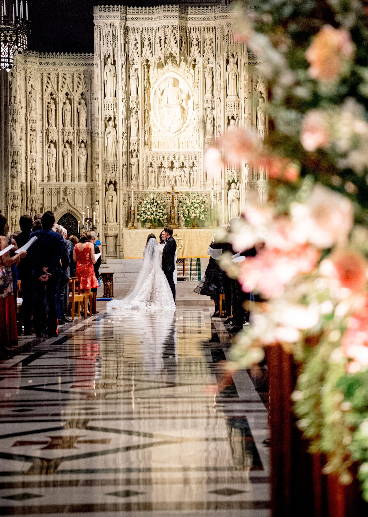 At the Washington National Cathedral in Washington D.C., this enchanting moment was expertly captured by the talented photographer Twah Dougherty, with Current Films as the videographers. Here, the bride and groom stand at the altar of the magnificent church, exchanging vows during their wedding ceremony. The intricate carvings of the church create a stunning backdrop, while the foreground features a beautiful array of flowers and guests seated on either side of the aisle. The glossy marble flooring adds to the elegance of the scene, making it a truly memorable occasion.