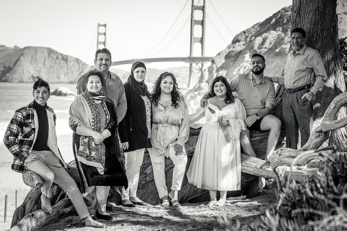 Black and white image of a large family posing for photos on baker beach with the golden gate bridge in the background