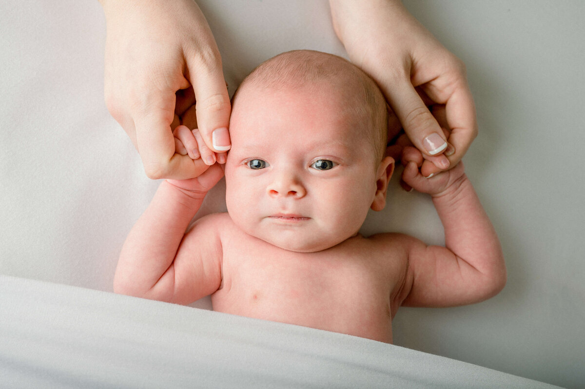 A newborn baby laying on a white sheet with a white blanket, holding mom's hands and looking at the camera.