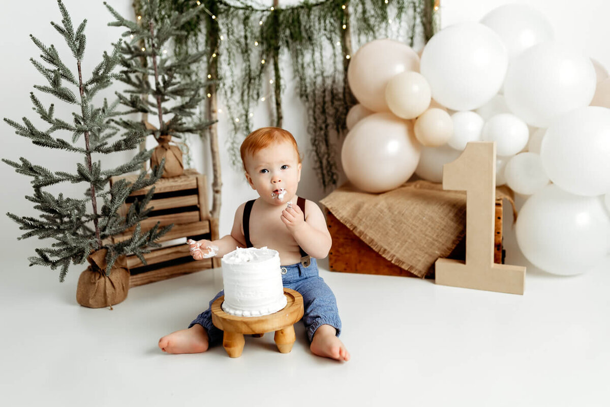 A smiling baby with red hair wearing a brown overalls and white shirt sits in front of a festive background with balloons and greenery. Twinkling lights add a warm ambiance.