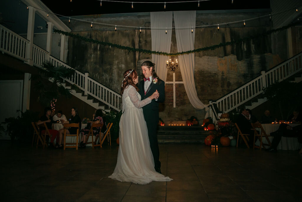 A bride and groom share their first dance in a warmly lit venue with draped fabric, greenery, and pumpkins decorating the space. Guests are seated at tables in the background, watching the couple.