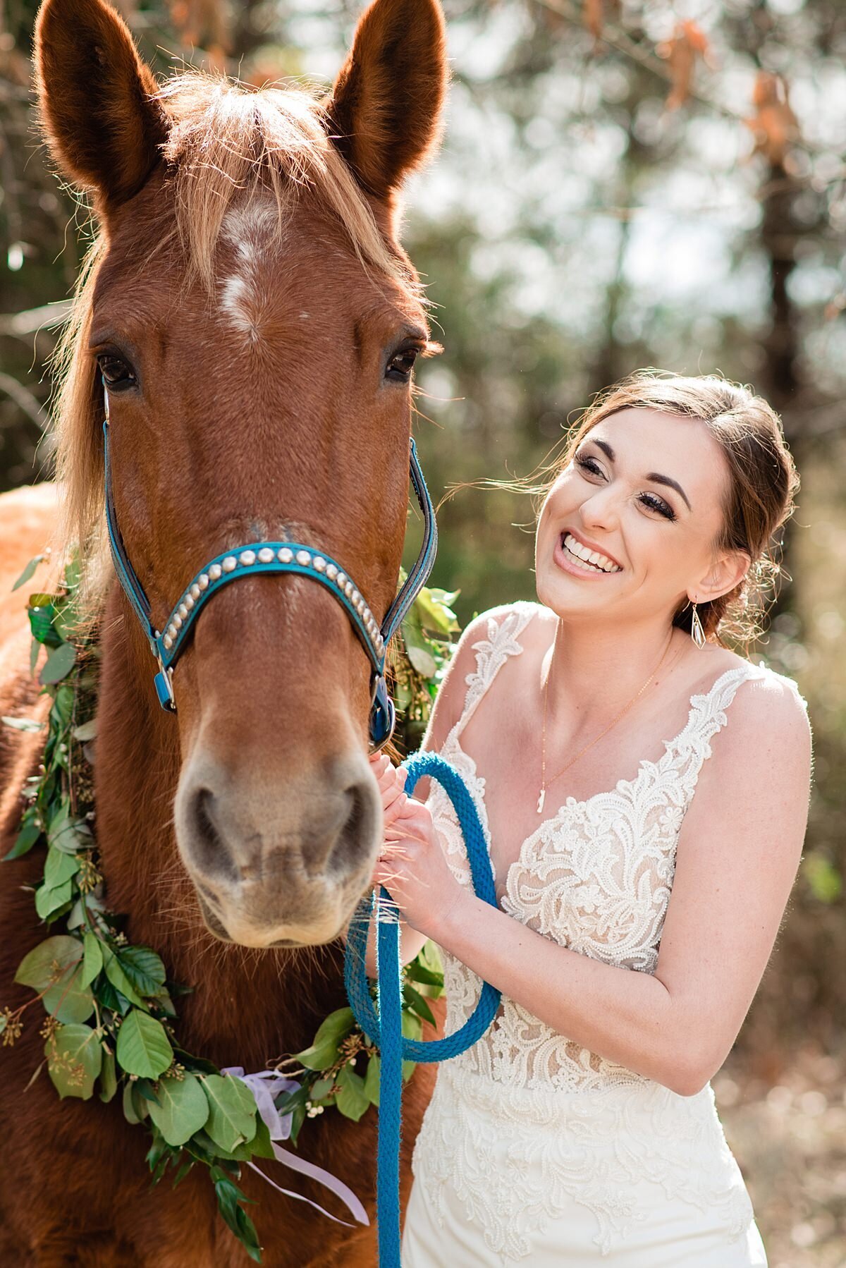 A bride wearing a lace wedding dress with a plunging neckline smiles up at her brown thoroughbred horse who is wearing a teal harness and a wreath of greenery at Grace Valley Farm.
