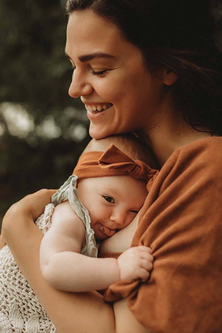 A beautiful mother holds her baby under her chin while she smiles with her eyes closed. The baby sticks her tongue out while hugging her mom.