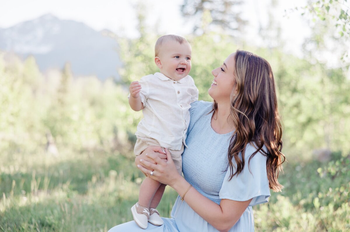 A mom in a blue dress plays with her baby boy in her lap in a white shirt in a mountain meadow for a Breckenridge Family Photographer