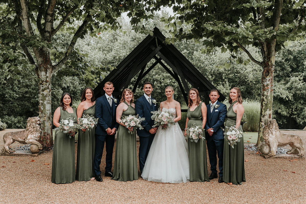 Bride and Groom having their photo taken with bridesmaids and groomsmen in front of black wooden archway at Bury Court Farnham