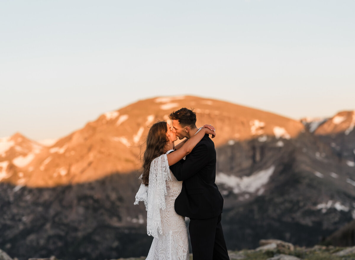 A bride and groom kiss in the sunrise with a mountain behind them