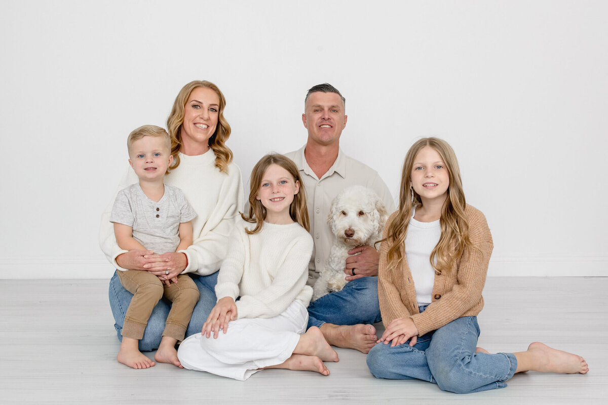A family of five wearing white, blue, and brown sitting together on a grey floor with their white, fluffy dog, smiling at the camera in a white studio.