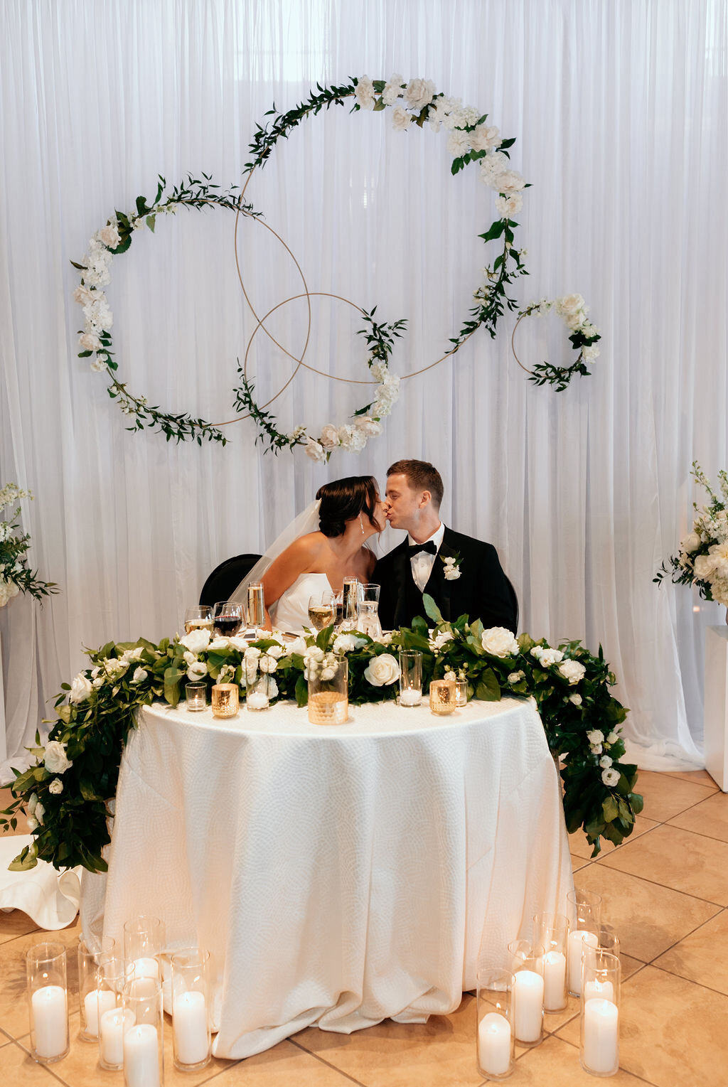 Bride and groom at their sweetheart table during a spring wedding at the Palm Event Center in Pleasanton, CA. The elegant setup features a backdrop of white drapery and gold metal wreaths adorned with white flowers.