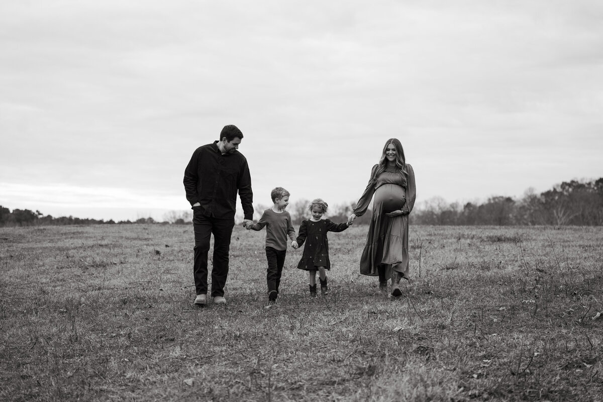 Black and white maternity photo of a family walking hand-in-hand through a field—capturing connection, anticipation, and timeless joy.