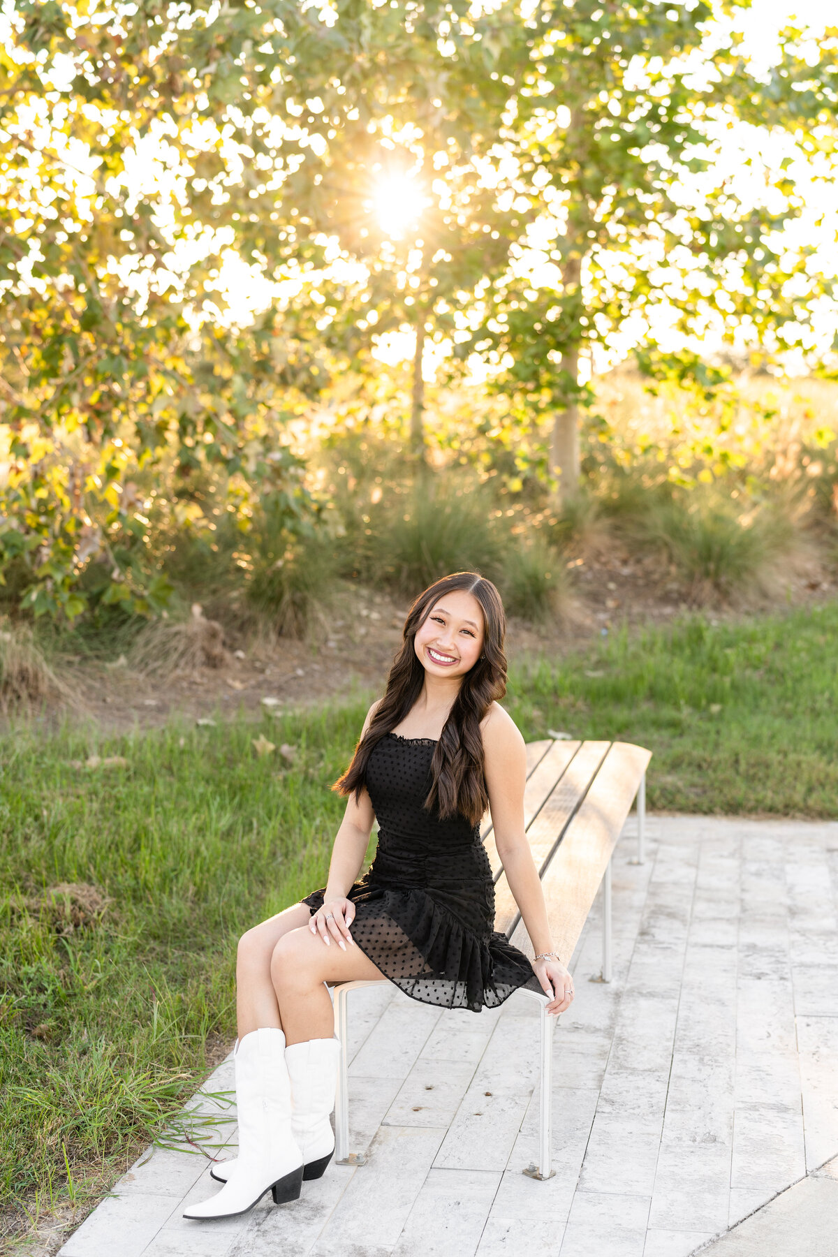 Houston High School senior girl wearing black dress and smiling while sitting on bench surrounded by nature at sunset at Josey Lake Park