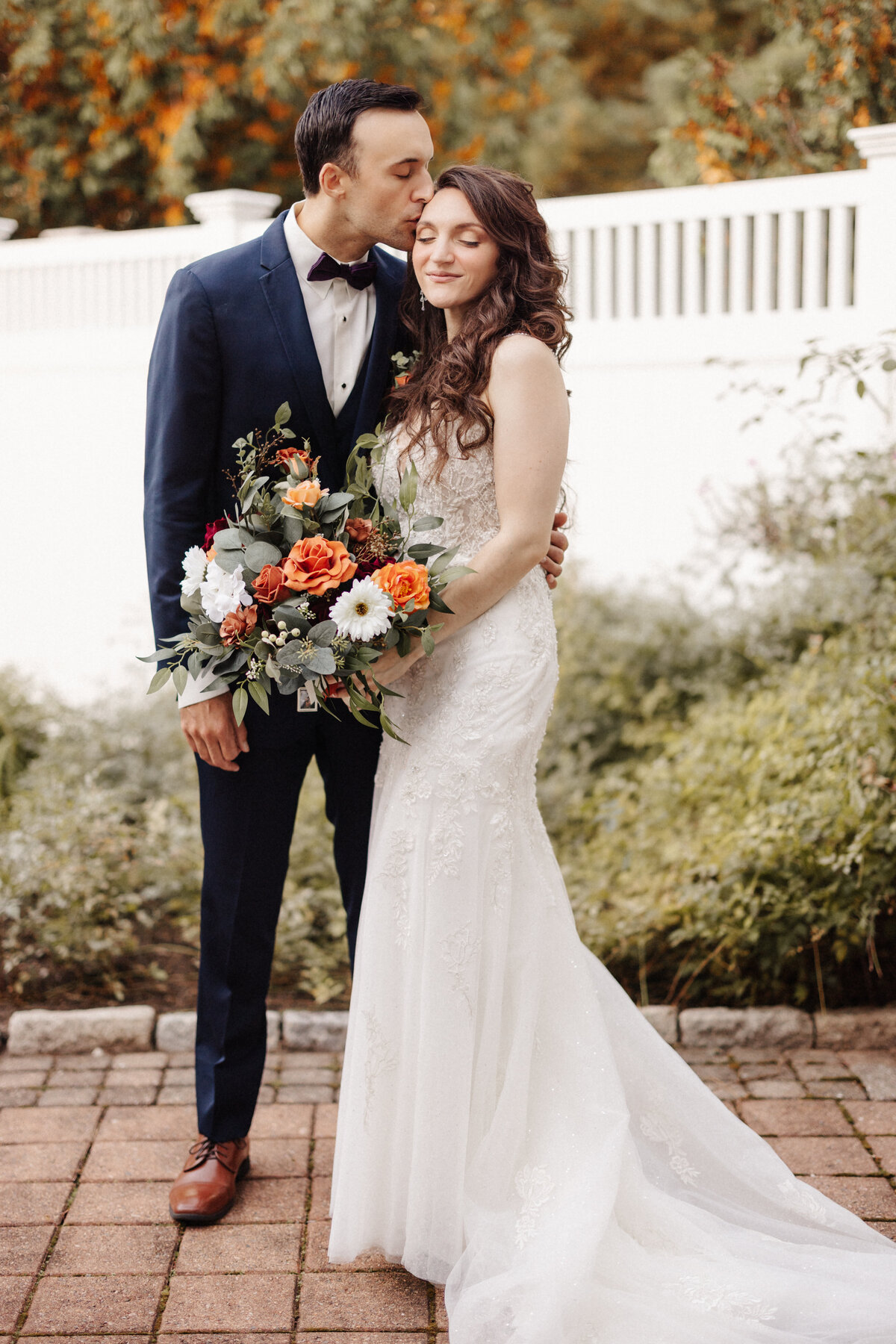 Romantic wedding portrait of a bride holding a fall bouquet as the groom kisses her forehead outdoors.