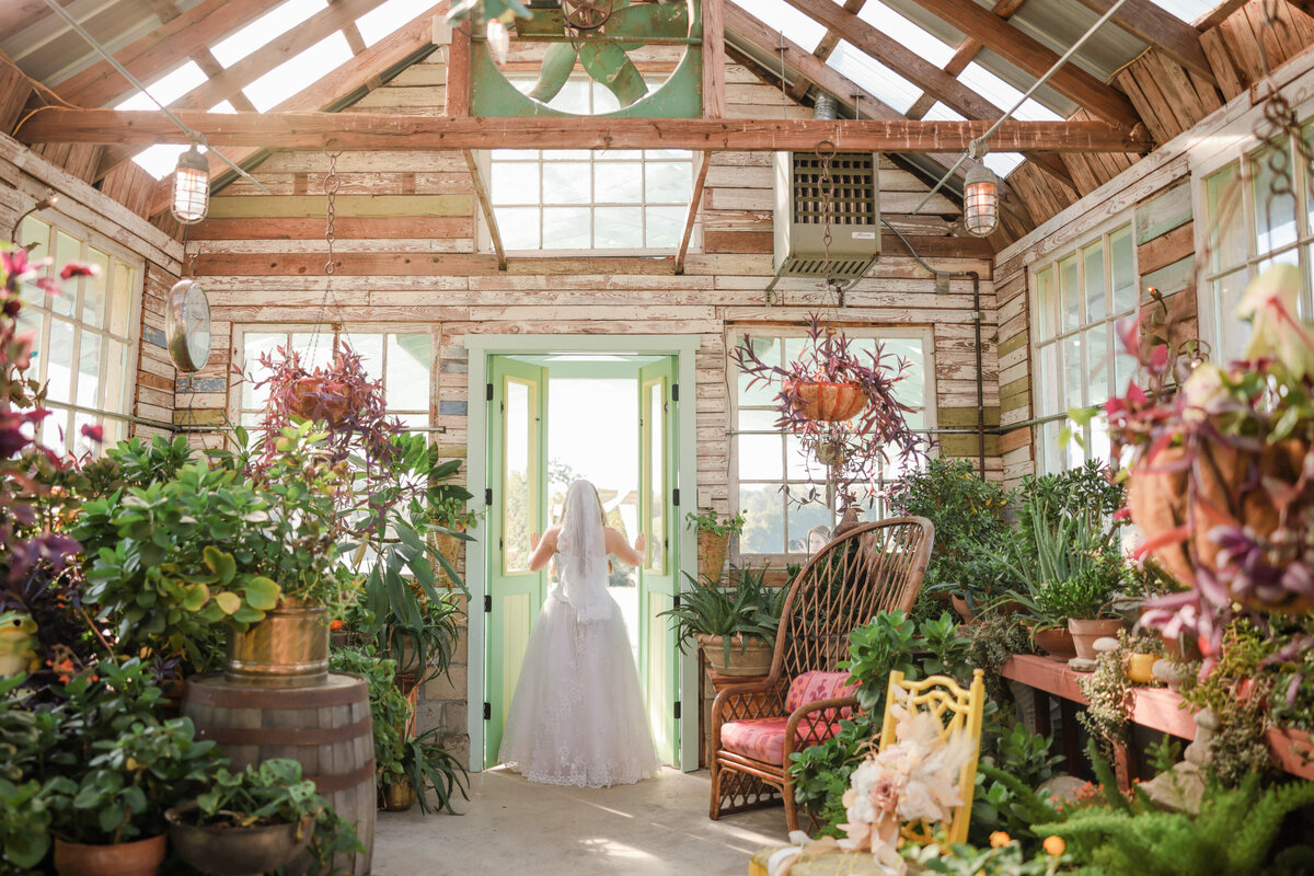 Bride stands at greenhouse doors showing off her beautiful gown and veil.