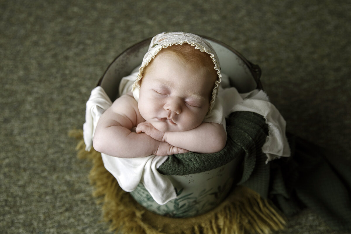 Portrait of a baby in a bucket posed with its chin on her hands and wearing a cream colored bonnet