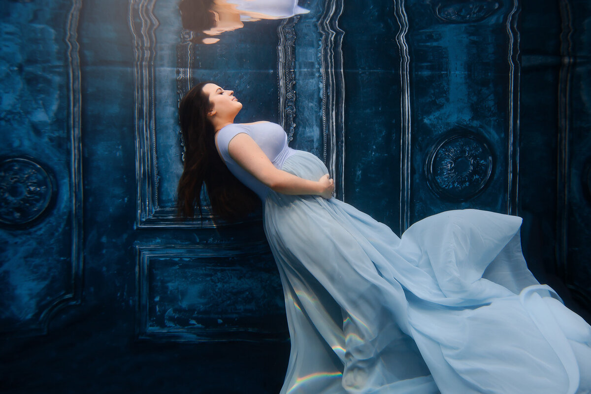 A serene underwater maternity photo showcasing a woman in a flowing pale blue gown against a richly detailed, deep blue backdrop. The ethereal ambiance, enhanced by soft light and reflections, creates a mesmerizing Dallas underwater maternity photoshoot experience.