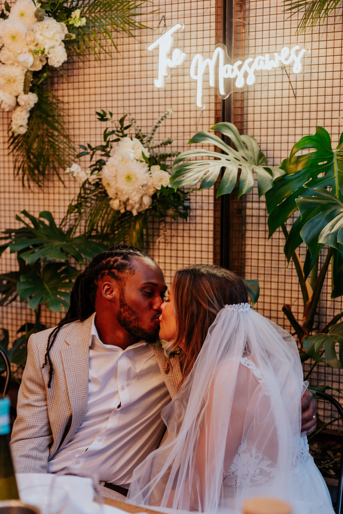 Bride and groom kiss infront of a backdrop of plants and a neon sign that says 'The Massawes'