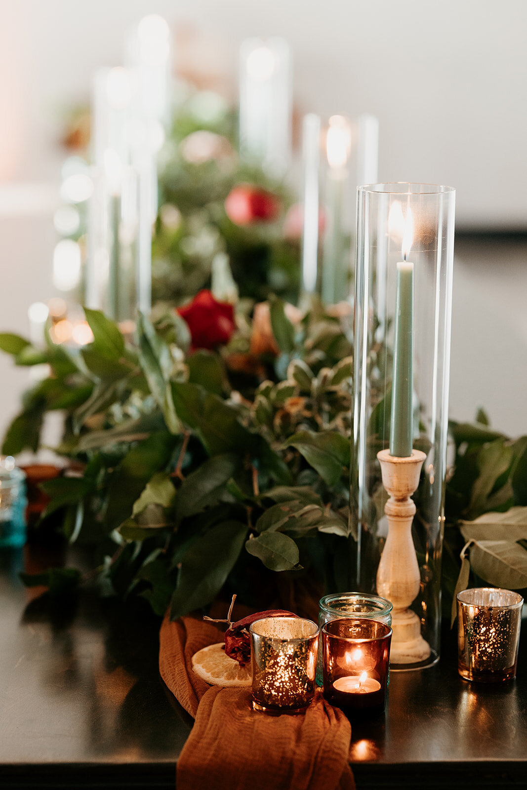 An elegant tablescape with greenery, roses, and tall green candles in glass holders, complemented by smaller candles and warm accents.