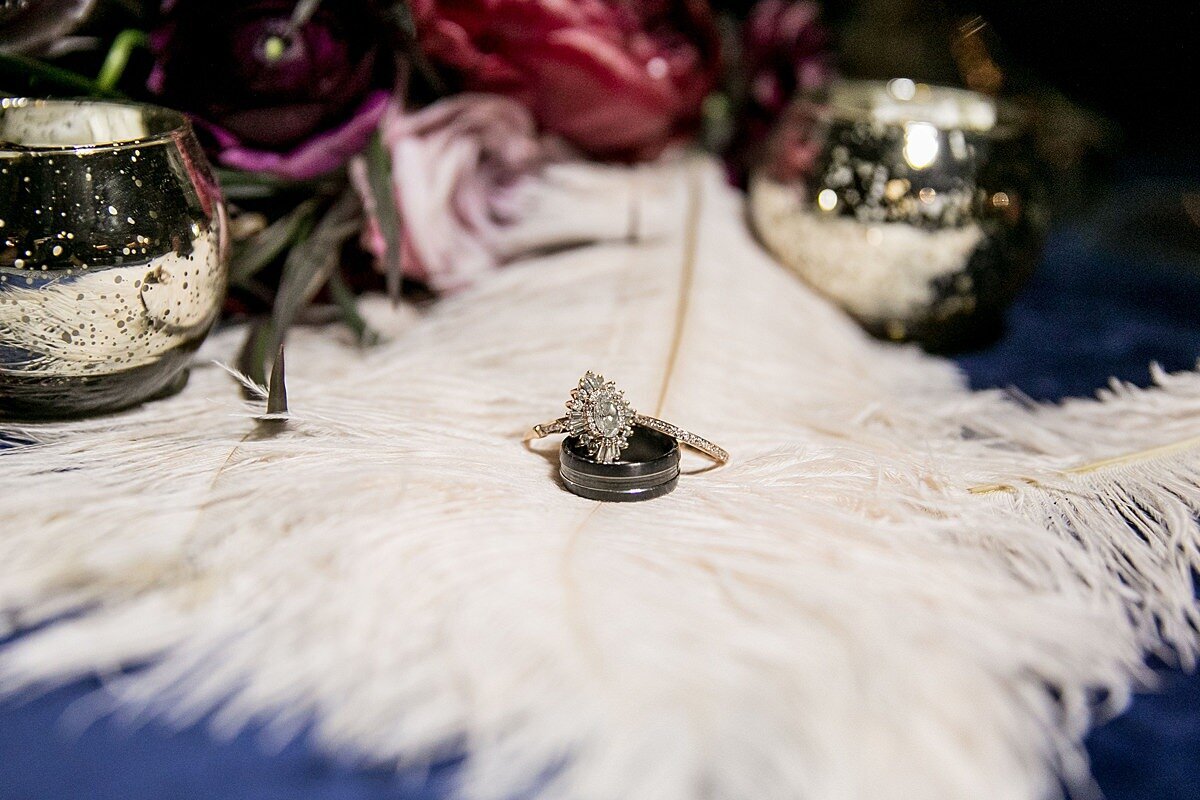 A vintage diamond engagement ring and wedding bands sit on top of a white ostrich feather plume on the navy blue velvet table cloth reception table surrounded by burgundy peonies and gold votive candles.