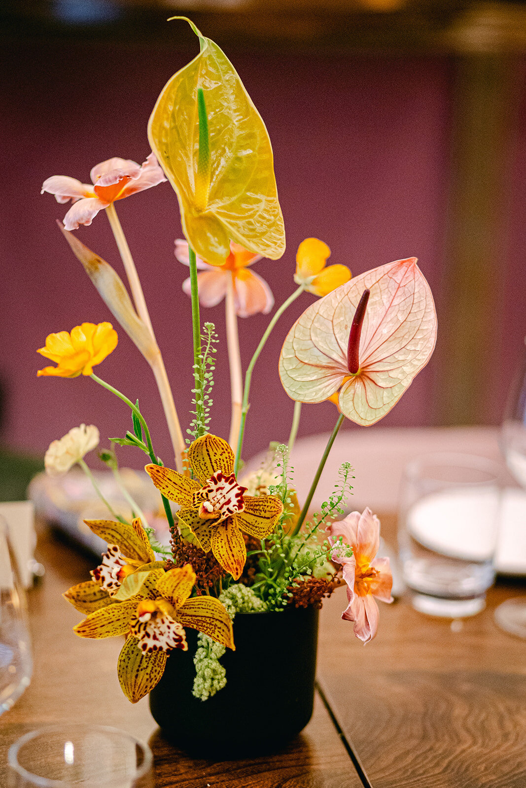 Modern floral centerpiece with tropical anthuriums, orchids, and golden-hued flowers in a black vase