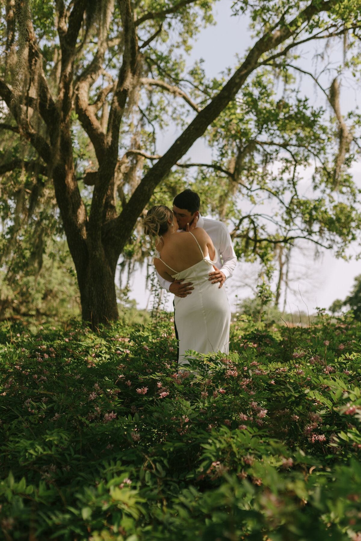 A wedding couple kissing while standing in the middle of a patch of wildflowers underneath the moss draped oak trees of Louisiana. 