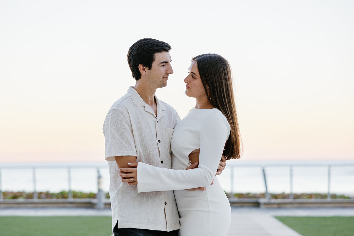 Rebecca and Graysen, dressed in sophisticated white outfits, share an intimate moment by the ocean, with the stunning backdrop of Marriott Hotel Fort Lauderdale enhancing their engagement photos.