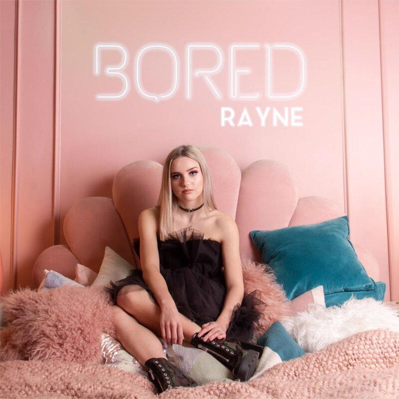 Single Cover Artist Rayne Title Bored singer sitting on pink bed surrounded with pillow word Bored in neon white lights on wall behind her