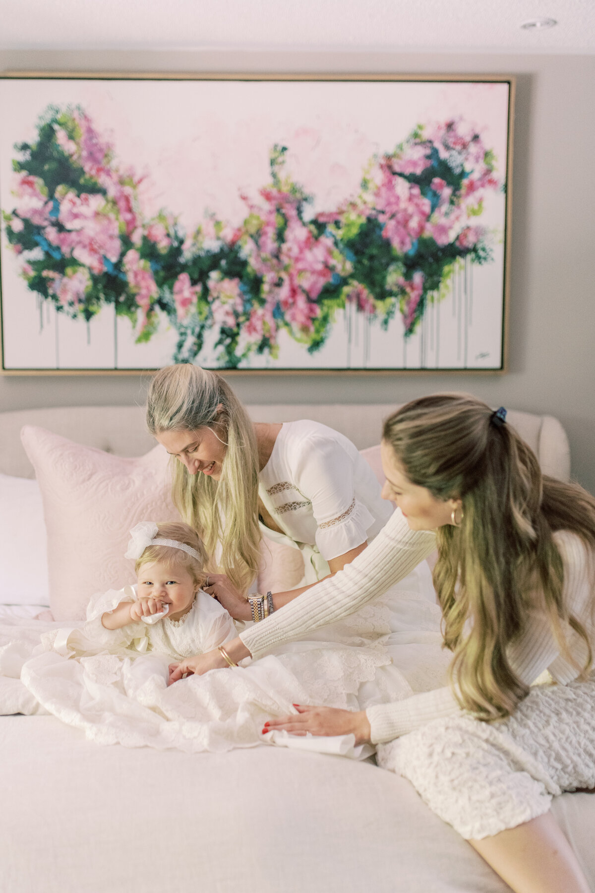 Serene portrait of mother and godmother cradling baby on soft bedding, mirror reflecting floral tones.