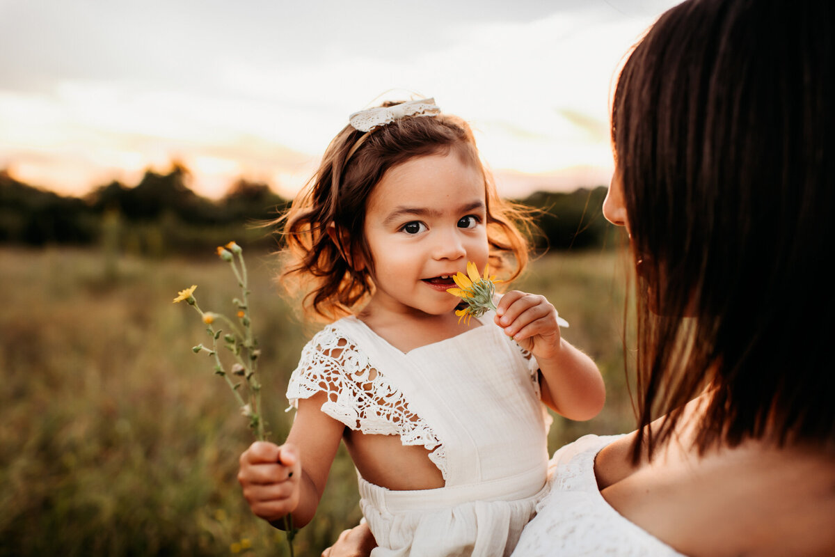 Mom holding little girl, who is smelling flowers.