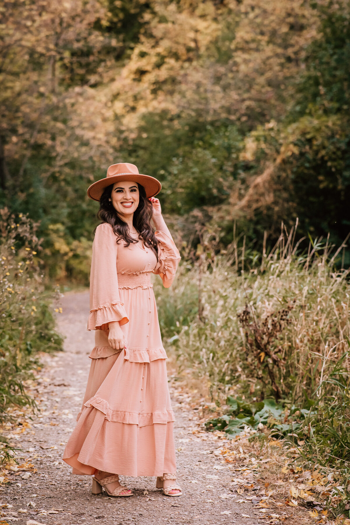 A woman in a flowing peach-colored dress and matching hat stands on a nature trail, smiling warmly at the camera, with autumn foliage in the background.