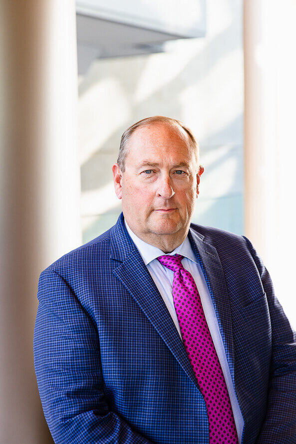 Man in blue jacket and pink tie stand in front of column for his photo.