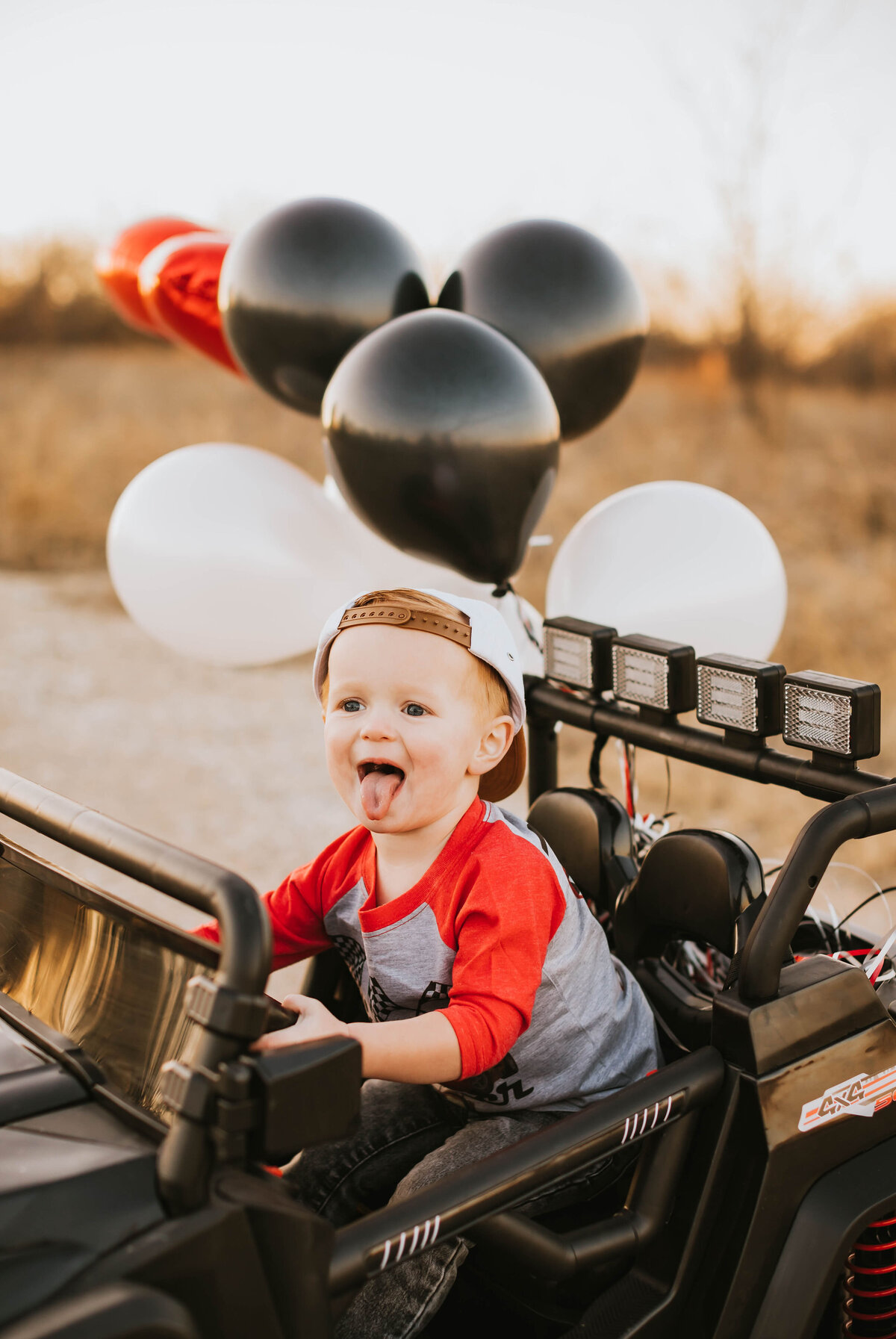 toddler boy sticking his tongue out in a black jeep with balloons tied to it