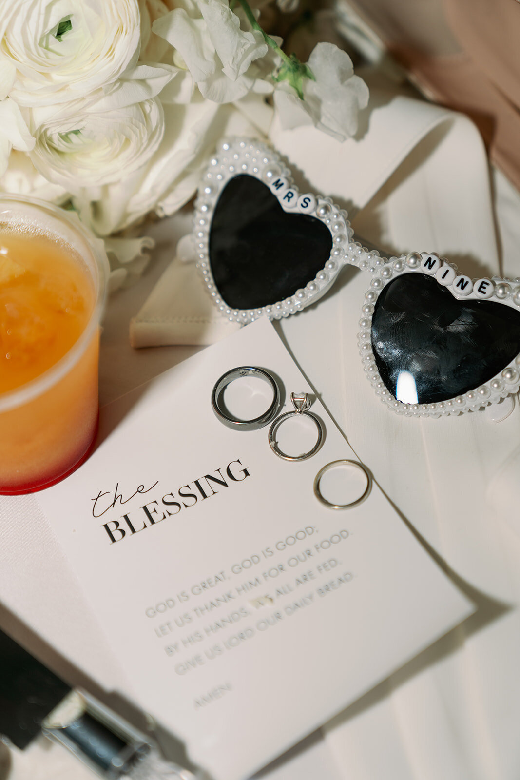 Wedding details flatlay featuring heart sunglasses, the blessing program, and rings at The Morris Estate.