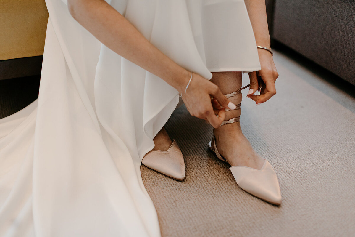 The bride, wearing a flowing white dress, is tying the ribbon on her pointed satin shoes, preparing for the ceremony.