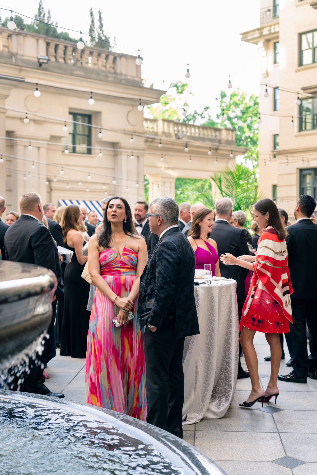A formal outdoor event with people mingling. A woman in a colorful dress and a man in a dark suit stand near a fountain. More guests are gathered around tables, and string lights are overhead. Buildings and greenery are visible in the background.